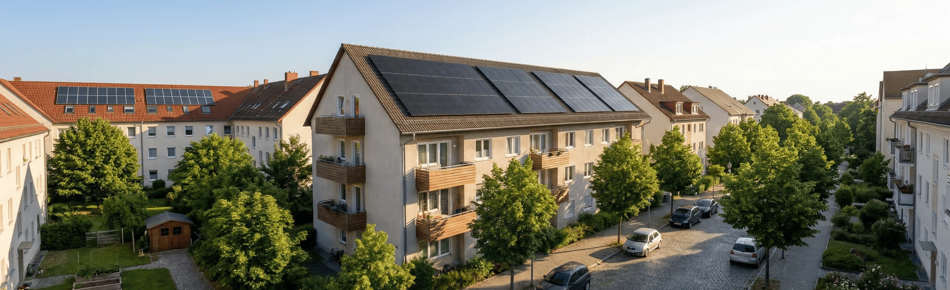 Wohngebäude mit Solarpaneelen auf den Dächern, umgeben von grünen Bäumen und parkenden Autos an einer Kopfsteinpflasterstraße.